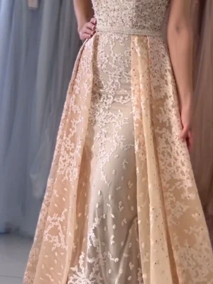 Champagnefärgad långklänning med spets - Elegant långklänning i champagnefärg med vackra spetsdetaljer och broderade blommor. Klänningen har ärmlös design, v-ringad rygg och ett lager av transparent tyg över en slät underklänning. Perfekt för speciella tillfällen.