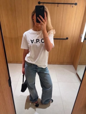 Vit A.P.C. t-shirt med tryck - Säljer en klassisk vit t-shirt från A.P.C. med svart logotyptryck framtill. T-shirten har rund halsringning och korta ärmar. Tillverkad i mjuk bomull för en skön och avslappnad look. Perfekt att styla med jeans eller kjol.