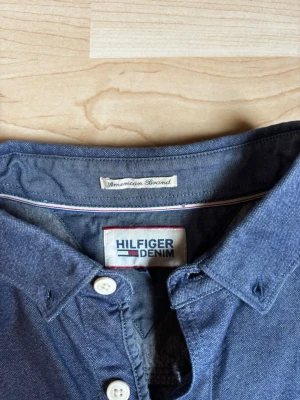 Blå skjorta från Hilfiger Denim - Snygg blå skjorta från Hilfiger Denim med klassisk krage och vita knappar. Skjortan har lång ärm och liten logga på bröstet. Perfekt för dig som gillar stilrena och tidlösa plagg. Kan stryka skjortan vid önskemål om det:)