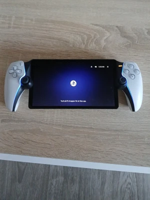 PlayStation Portal handhållen konsol - PlayStation Portal är en handhållen spelkonsol från Sony med stor svart pekskärm och vita kontroller på sidorna, inspirerade av DualSense-designen. Konsolen har blå LED-detaljer och ergonomiska handtag för bästa spelupplevelse.