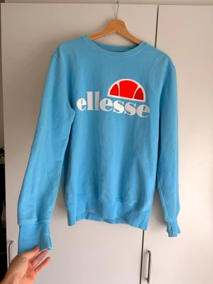 Blå sweatshirt från Ellesse - Snygg ljusblå sweatshirt från Ellesse med stort vitt och rött logotryck på bröstet. Knappt använd! Otroligt fin färg men den är tyvärr  lite för stor för mig😔. Tröjan har rund halsringning, ribbade muddar vid ärmslut och nederkant samt en avslappnad passform. 