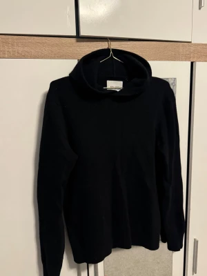 Stickat vailent hoodie - Mysig svart stickad polotröja från Vaillent i mjuk merinoull. Tröjan har lång ärm och hög polokrage, perfekt för kyliga dagar. Enkel och stilren design som passar till det mesta.