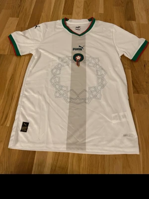 Vit Puma fotbollströja Marocko - Snygg vit fotbollströja från Puma med Marockos emblem på bröstet. Tröjan har gröna och röda detaljer vid krage och ärmslut samt ett diskret mönster framtill. Tillverkad i polyester med korta ärmar och sportig passform.