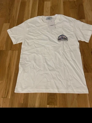 Vit Casablanca t-shirt med tryck - Säljer en vit t-shirt från Casablanca med färgglatt tryck på bröstet och stort grafiskt motiv på ryggen. T-shirten har korta ärmar och rund hals. Perfekt för dig som gillar stilrena plagg med unika detaljer.
