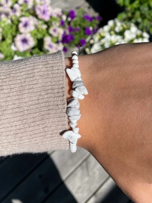 Vitt pärlarmband med stenar - Snyggt armband med vita pärlor och oregelbundna vita stenar med grå marmorering. Armbandet har små guldfärgade detaljer och justerbar knytning i vitt snöre. Perfekt accessoar för en trendig och stilren look.