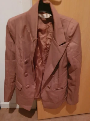 Kavaj - Snygg brun kavaj med dubbelknäppning och breda slag. Kavajen har två framfickor med lock och är helfodrad. Perfekt för dig som vill ha en trendig och stilren look. Passar till många olika outfits. Det är i kinesiskt storlek men motsvarar svensk storlek 34/36