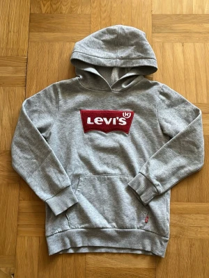 Grå hoodie från Levi's med röd logga - Snygg grå hoodie från Levi's med stor röd broderad logga på bröstet. Klassisk känguruficka framtill, ribbade muddar och mjukt material som känns skönt mot huden. Perfekt för en avslappnad och trendig stil. köpt i usa passar runt 135-140cm