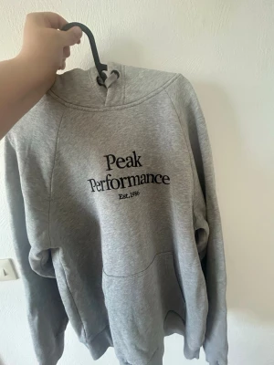 Grå hoodie Peak Performance - Klassisk grå hoodie från Peak Performance med svart logga och text på bröstet. Hoodien har huva, känguruficka och långa ärmar. Nästan aldrig använd & är som nyskick. Djur & rökfritt hem.