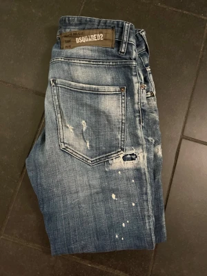 Blå slitna jeans från Dsquared2 - Säljer ett par blå jeans från Dsquared2 med coola slitningar, färgstänk och tvättad look. Jeansen har klassisk femficksmodell och raka ben. Perfekta för dig som gillar en avslappnad och trendig stil. De har inte för mycket slitningar för dig som är rädd för de. Dsquard2 har en annan storlek än ”vanliga” jeans märken så kolla upp din speciella storlek på dsquard2 eller fråga mig kan hjälpa lite. Skick är svårt att säga eftersom jeansen redan har speciella slitningar