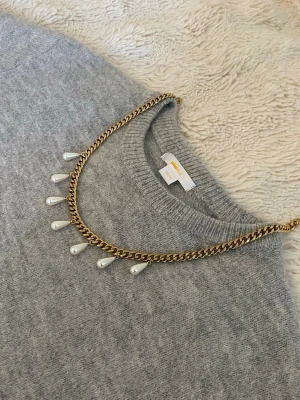 Guldhalsband med droppformade pärlor - Snyggt halsband i guldfärgad rostfritt med droppformade vita pärlor som hänger längs kedjan. Kedjan har en chunky känsla och pärlorna ger en elegant touch. Perfekt för att lyfta vilken outfit som helst med en trendig och stilren look.