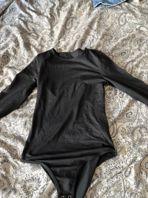 Svart långärmad body XS - En enkel och stilren svart långärmad body i storlek XS. Tillverkad i mjukt och stretchigt material med 95% polyester och 5% elastan. Rund halsringning och tryckknappar i grenen för enkel på- och avklädning. Perfekt att matcha med jeans eller kjol.