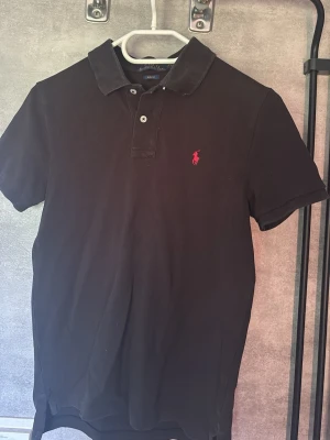 Ralph Lauren Piké - Klassisk svart pikétröja från Polo Ralph Lauren i slim fit-modell. Tröjan har korta ärmar, krage och knappar framtill samt den ikoniska röda loggan broderad på bröstet. Perfekt för en clean och stilren look.  Storlek: 14-16yr/XS/ mindre S.