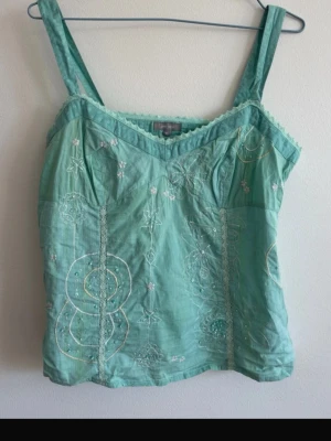 Vintage Mintgrön broderad linnetopp - Söt mintgrön linnetopp med breda axelband och v-ringad hals. Toppen har fina broderade detaljer i vitt och ljusgult samt spets längs urringningen. Perfekt för varma dagar och ger en fräsch look.