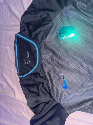 Svart Nike fotbollströja med blå detaljer - Snygg svart fotbollströja från Nike med blå logga och emblem på bröstet. Tröjan har ett diskret rutmönster framtill och blå kantband vid hals och ärmslut. Tillverkad i lätt och ventilerande material, perfekt för träning eller match.