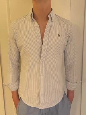 Vitblå randig skjorta från Ralph Lauren - Klassisk vit och ljusblå randig skjorta från Ralph Lauren med button-down krage och broderad logga på bröstet. Skjortan har långa ärmar, normal passform och stängs med knappar framtill. Perfekt för sommaren. Den passar personer som är unge för 170 cm. Fråga gärna om mått och pris kan sänkas vid snabb affär 