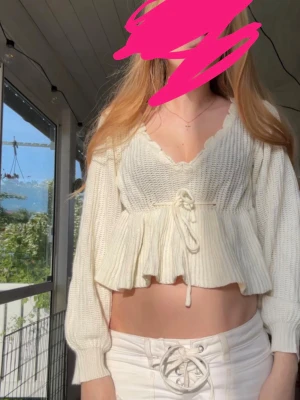 Vit stickad topp med volang - Säljer en söt vit stickad topp med volangkant och knytband i midjan. Tröjan har djup v-ringning, långa ärmar och ribbade muddar. Perfekt för dig som gillar romantisk och trendig stil.