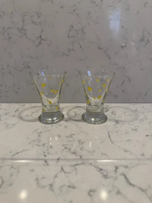 Två små glas med gula citroner  - Säljer två små glas med utsvängd form och dekorativa citroner prickar runt om. Perfekta för shots eller små drinkar. Glasen är genomskinliga och har en stadig fot. Snygga att ha framme i köket eller till festen.