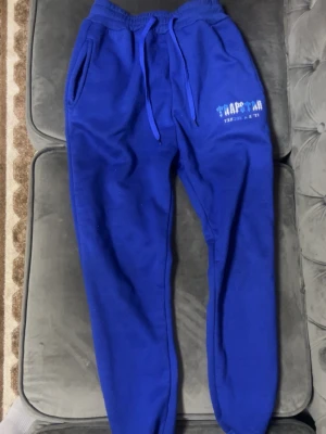 Blå mjukisbyxor från Trapstar - Säljer ett par blå mjukisbyxor från Trapstar med tryckt logga på vänster sida. Byxorna har dragsko i midjan, två sidofickor och muddar vid bensluten. Perfekta för chill dagar och streetwear-stil. Pris kan diskuteras 