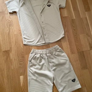 Beige prada  - Säljer ett beige loungewear som är märket Prada med kortärmad skjorta och matchande shorts. har en diskret rutstruktur, knappstängning på skjortan och elastisk midja på shortsen. Båda delarna har Pradas ikoniska trekantslogga. Perfekt för chill dagar hemma.
