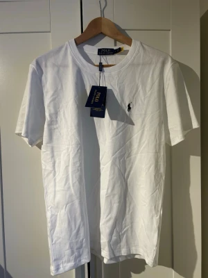 Vit t-shirt från Polo Ralph Lauren - Klassisk vit t-shirt från Polo Ralph Lauren med rund halsringning och korta ärmar. T-shirten har den ikoniska svarta polospelaren broderad på bröstet och är tillverkad i mjuk bomull. Perfekt för en stilren och enkel look. Helt ny priset kan diskuteras 