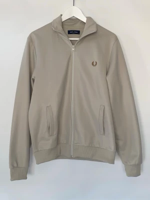  ziptröja Fred Perry - Säljer denna snygga beige ziptröja från Fred Perry med klassisk laurel-logga broderad i guld på bröstet. I väldigt bra skick i storlek M