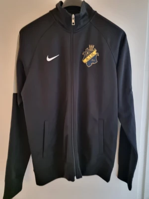 Svart AIK träningsjacka från Nike - Snygg svart träningsjacka från Nike med AIK:s klubbmärke på bröstet. Jackan har hel dragkedja framtill, ståkrage och två sidofickor. Perfekt för dig som vill representera AIK både på och utanför planen.