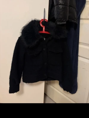 Svart teddyjacka med fuskpäls-krage - Mysig svart teddyjacka med stor fluffig fuskpäls-krage och knäppning framtill. Använt fåtal gånger ,Nypris 439 kr! Tveka inte att höra av er vid frågor eller funderingar💗🥰