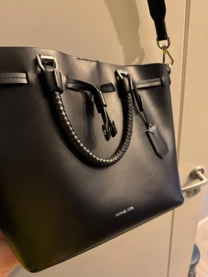 Svart handväska från Michael Kors - Stilren svart handväska i slätt konstläder från Michael Kors med flätade handtag, silverfärgade metalldetaljer och avtagbar axelrem. Väskan har dekorativa remmar och metallfötter undertill för extra skydd. Perfekt för dig som gillar klassisk och modern design.