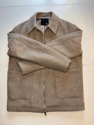 Beige overshirt från 157 - Stilren beige overshirt från 157 i mjukt ull-liknande material. Jackan har klassisk krage, två stora framfickor och dragkedja framtill. Perfekt lager-på-lager-plagg med minimalistisk design och raka linjer. Storlek XS (passar även S). Skick 9,5/10 använd ca 2-3 gånger.