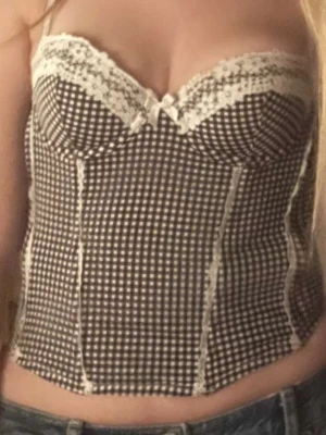 Rutig korsettopp med spetsdetaljer - Supersöt korsettopp med svartvitt rutigt mönster och spetsdetaljer upptill. Dekorerad med en liten rosett i mitten och har markerade sömmar för en snygg siluett. Perfekt för dig som vill ha en trendig och feminin look.