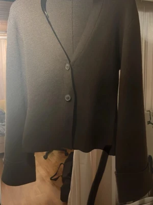 Brun kofta från Gina Tricot - Mörkbrun kofta från Gina Tricot med v-ringning och två knappar framtill. Modellen är croppad med långa ärmar och breda muddar. Materialet är mjukt och stickat, perfekt för lager-på-lager-stil.