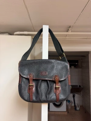 Mulberry axelväska – äkta läder, Made in England - Säljer en klassisk Mulberry axelväska i mörkbrunt/gråtonat läder med bruna läderdetaljer. Väskan är i äkta läder av hög kvalitet och tillverkad i England, vilket syns på märkningen inuti.  ✨ Detaljer: 	•	Original Mulberry 	•	Äkta läder 	•	Justerbar axelrem 	•	Lock med remmar/spännen 	•	Flera innerfack – praktisk och rymlig 	•	Klassisk tidlös modell som passar både vardag och jobb  📌 Skick: Använd men i gott skick. Naturlig patina och vissa bruksspår finns (se bilder), vilket är normalt för Mulbe