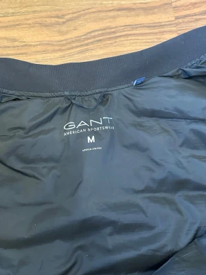 Svart dunväst från GANT - Snygg svart dunväst från GANT med quiltat mönster och dragkedja framtill. Västen har två fickor med dragkedja och en diskret logga på bröstet. Perfekt för lager på lager och sportig stil. Tillverkad i lätt och smidigt material. Skicket på västen är 8/10. Priset kan diskuteras 