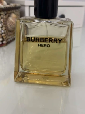 Burberry Hero Eau de Parfum (70ml-80ml) - Burberry Hero är en lyxig parfym med en stilren, fyrkantig glasflaska och svart kork med Burberrys logga på toppen. Doften har en gyllene färg och flaskan utstrålar modern elegans. Perfekt för dig som vill ha en ikonisk och trendig doft i samlingen. Följ oss för 12% rabatt 