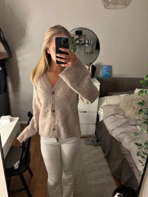 Beige kofta med knappar - Mysig beige kofta med v-ringning och stora knappar framtill. Koftan har en avslappnad passform, långa ärmar och är stickad i ett mjukt material. Perfekt att slänga på sig över en topp för en chill look.