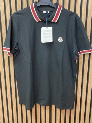 Svart pikétröja  - Svart pikétröja  med klassisk krage och röda samt vita ränder på krage och ärmslut. Tröjan har korta ärmar, knappar framtill och  broderad på bröstet. Tillverkad i bomull för en bekväm känsla.