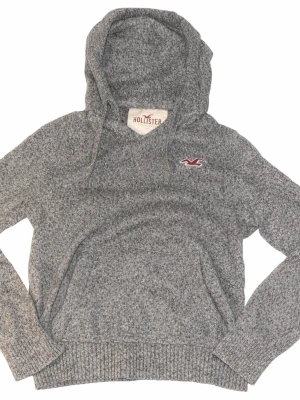 Grå hoodie från Hollister - Mysig gråmelerad hoodie från Hollister med huva och dragsnören. Tröjan har lång ärm, ribbade muddar och Hollisters klassiska logga i rött och vitt broderat på bröstet. Perfekt för chill dagar och enkel att matcha med jeans eller mjukisbyxor.