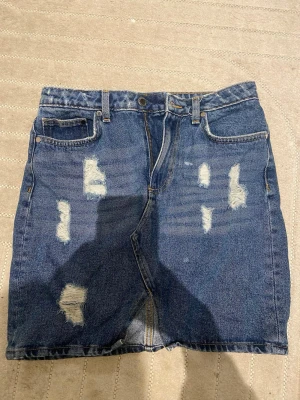 Blå jeanskjol med slitningar - Säljer en blå jeanskjol i klassisk modell med flera slitningar framtill för en trendig look. 