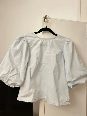 Ljusblå blus med puffärmar - Supersöt ljusblå blus från H&M med stora puffärmar och öppen rygg med knytband. Blusen har en lös passform och är tillverkad i ett lätt bomullstyg. Perfekt för dig som gillar trendiga och feminina detaljer.