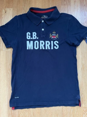 Marinblå piké från Morris Gents - Snygg marinblå pikétröja från Morris Gents med vit text 'G.B. MORRIS' och broderad logga på bröstet. Klassisk krage, korta ärmar och röda detaljer på insidan av kragen. Perfekt för en stilren och avslappnad look.