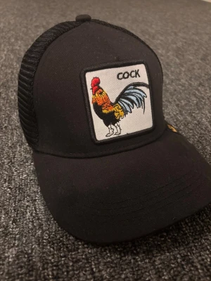 Svart goorin bros keps - Säljer en svart truckerkeps med mesh baktill och ett broderat märke framtill med en färgglad tupp och texten 'COCK'. Kepsen har böjd skärm och justerbar snapback. Perfekt för dig som vill sticka ut med en cool accessoar, från Goorin Bros. Äkta