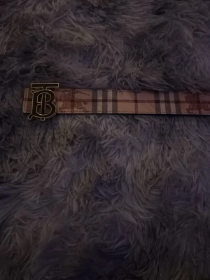 Rutig Burberry-bälte med BB-spänne - Snyggt bälte från Burberry med klassiskt rutigt mönster i beige, rött och svart. Bältet har ett stort metallspänne med BB-logga framtill. Perfekt accessoar för att lyfta din outfit med en lyxig touch.