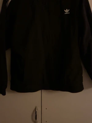 Svart hoodie från Adidas - Säljer en svart hoodie från Adidas med dragkedja och den klassiska vita loggan på bröstet. Hoodien har långa ärmar och en enkel, clean design som passar till det mesta. Perfekt för dig som gillar sportig och avslappnad stil.