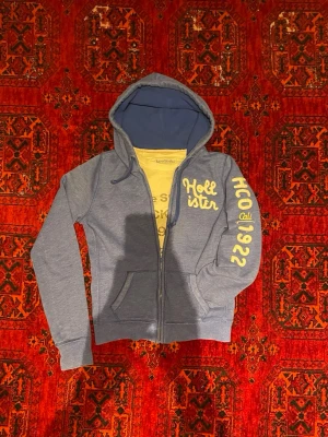 Blå Hollister Hoodie - Snygg blå zip hoodie från Hollister med vit text på bröstet och ärmen där det står 'HCO 1922'. Hoodien har två fickor fram, dragsko i huvan och mjukt material. Perfekt för en avslappnad och arcive stil. Undrar du något är det bara att skriva🙌