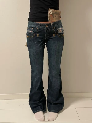 Mörkblå bootcut jeans  - Säljer ett par mörkblå bootcut jeans från med låg midja och snygga guldfärgade sömmar. Jeansen har dekorativa fickor både fram och bak samt detaljerade broderier. Perfekta för dig som gillar en retro och trendig look.