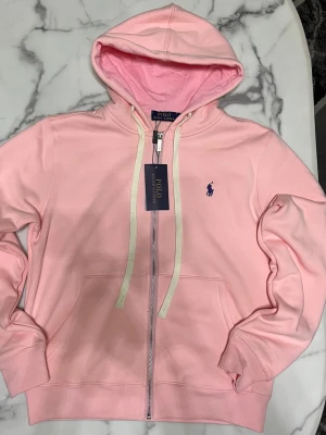 Polo Ralph Lauren rosa zip hoodie - Säljer en Polo Ralph Lauren zip hoodie i rosa. Plagget är helt nytt med original tags, aldrig använd.