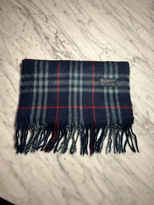 Mörkblå rutig halsduk från Burberry - Klassisk halsduk från Burberry i mörkblått med rutigt mönster i ljusblått och rött. Halsduken har fransar i ändarna och en vävd Burberry-etikett. Perfekt accessoar för att lyfta din outfit.