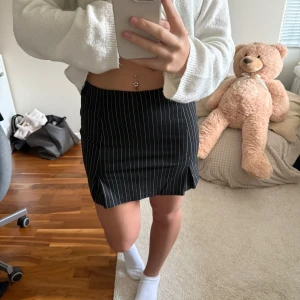 Svart minikjol med vita ränder - Snygg svart minikjol med tunna vita vertikala ränder och liten slits framtill. Kjolen har en tight passform och ger en trendig look. Perfekt att matcha med en croppad tröja eller stickad tröja 