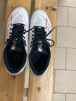 Vita Mizuno inomhusskor med röd detalj - Snygga vita inomhusskor från Mizuno med svarta snören och röda detaljer på sidorna. Skorna har meshpaneler för bra ventilation och en slitstark sula som passar perfekt för sporter inomhus. Klassisk låg modell med bekväm passform och svart innerfoder.