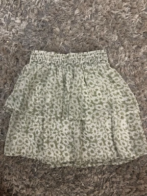 Grön blommig volangkjol - Supersöt kort kjol med volanger i ljusgrönt och vitt blommönster. Kjolen har resår i midjan för skön passform och är perfekt för vår och sommar. Materialet känns lätt och luftigt, vilket gör den enkel att styla med en crop top eller t-shirt.